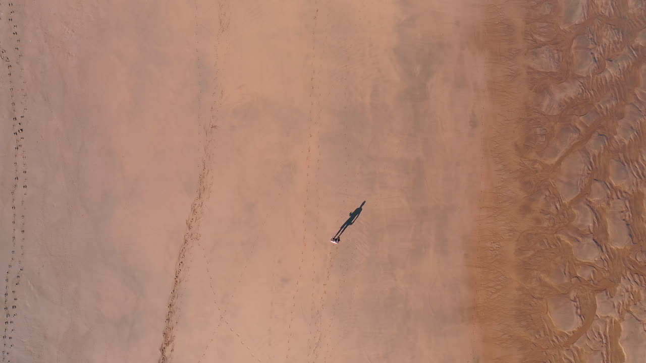 긴 그림자를 드리우는 썰물 동안 해변을 따라 걷는 사람의 하향식 공중 드론