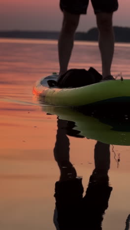 male silhouette reflecting in the river. lower part of a male legs standing on the sup board on the river at sunset. vertical video
