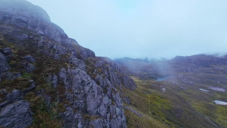 Cinematic-aerial-drone-shot-along-rocky-outcrop-through-the-fog-in-high-mountain-peaks-to-reveal-alpine-lake