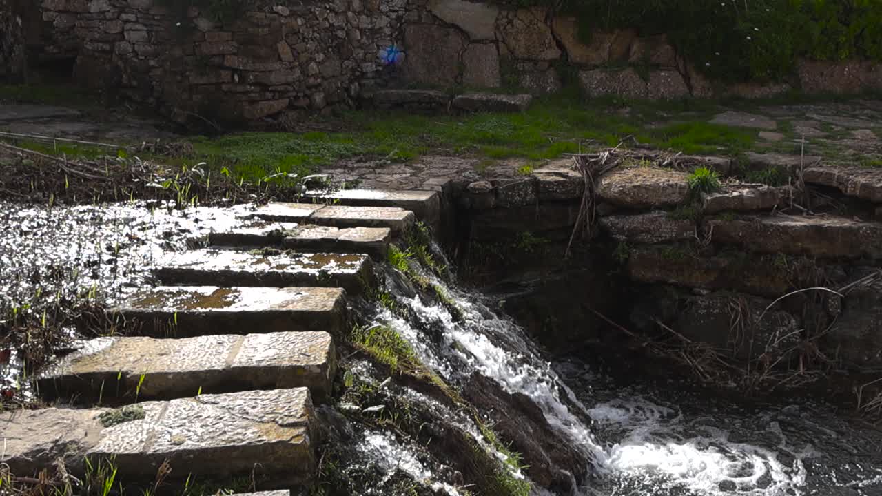 Sunny park stream flows over a small drop beside stone slab stepping crossing, creating bubbles and splashing water