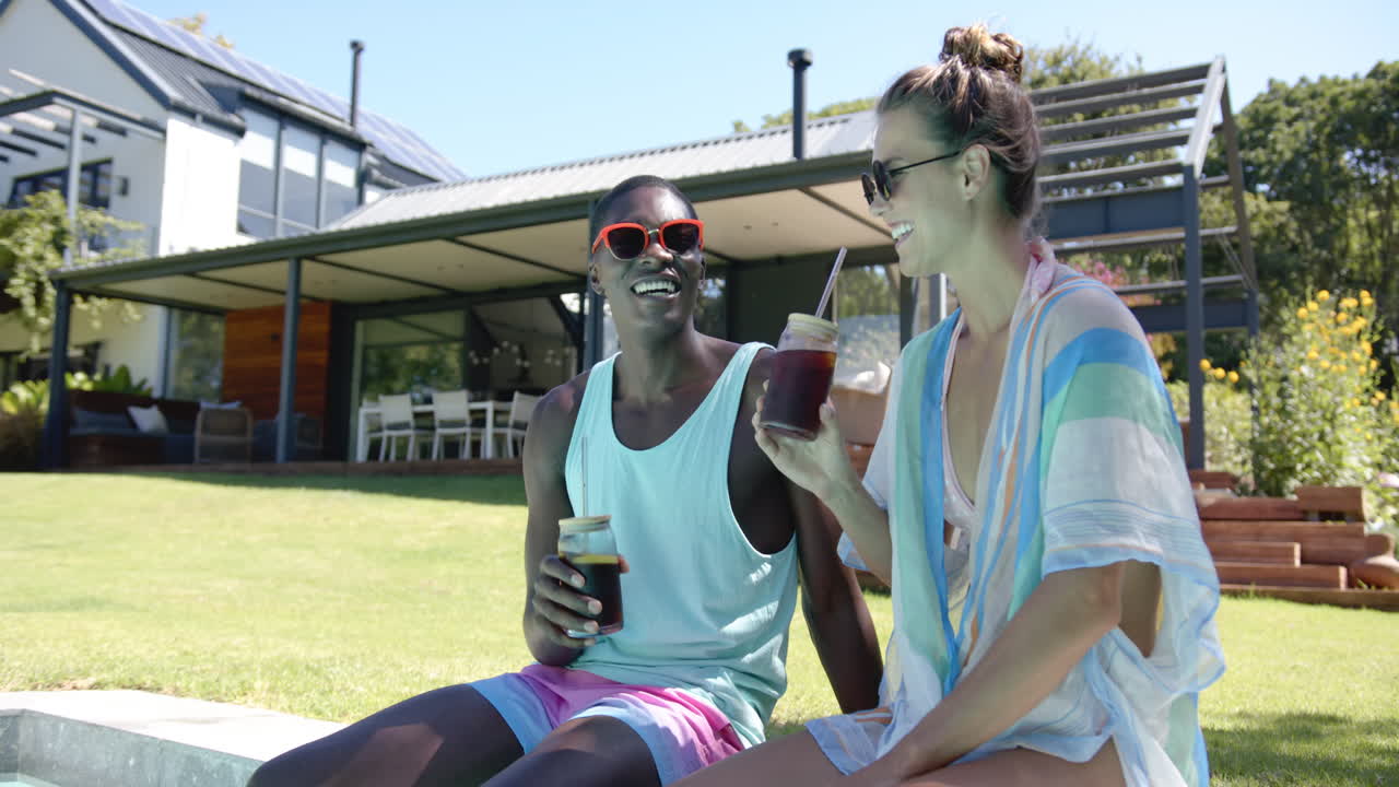 Relaxing by pool, diverse couple enjoying iced drinks and laughing together