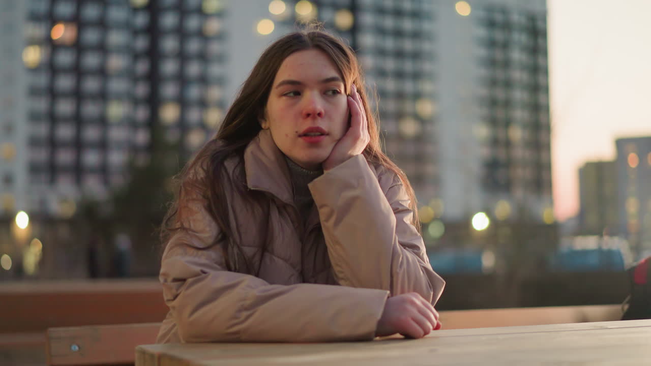 A close-up shot of a young woman wearing a peach jacket, lost in deep thought as she rests her cheek on her hand. Her eyes are closed, and the blurred urban background