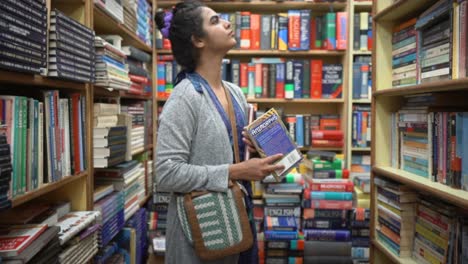 Asian-girl-standing,-holding-a-book-in-her-hand-and-thinking-by-looking-at-bookshelves,-Side-angle-shot