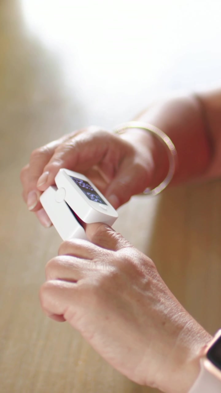 Vertical video: Glancing senior woman in red top reaching for oximeter at table, checking oxygen
