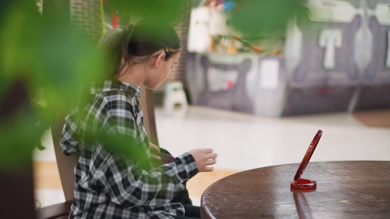una joven con camisa a cuadros se sienta en una mesa de madera, ajustando su cabello usando un espejo de madera, las plantas verdes en primer plano añaden un ambiente natural. la iluminación suave destaca su expresión tranquila y enfocada