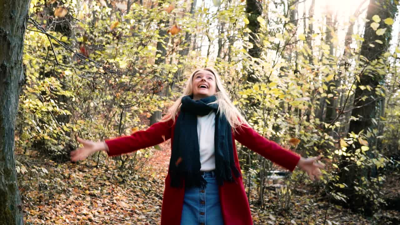 cámara lenta joven hermosa mujer arrojando hojas en el aire en medio de ti bosque de otoño naranja marrón mientras usa un abrigo rojo