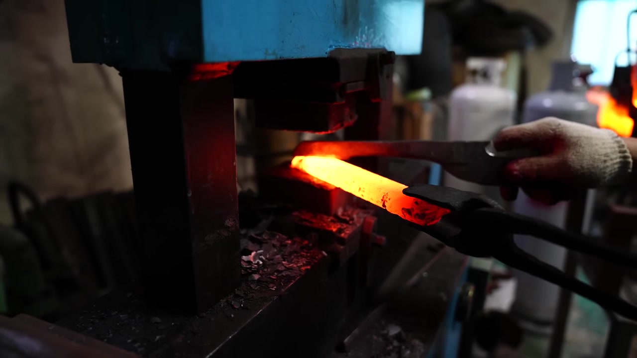 Tapering a glowing steel bar under a power hammer while gas cylinders stand in the workshop background