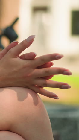 vista parcial de una mujer joven sentada al aire libre con las piernas cruzadas, frotando las manos pensativamente mientras disfruta de la cálida luz del sol, el fondo borroso presenta edificios y vegetación