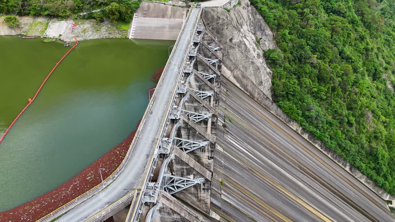 Aerial view of a large concrete dam with a road on top, reservoir on one side, and green hillside on the other