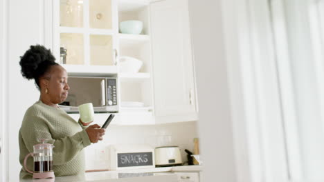 Happy-african-american-senior-woman-drinking-coffee,-using-smartphone-in-sunny-kitchen,-slow-motion