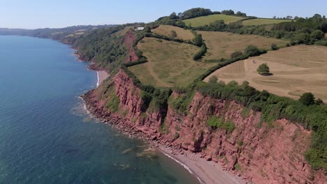 epic drone shot of red clay coastline, rocky shore, clear blue ocean water and green rural landscape on top