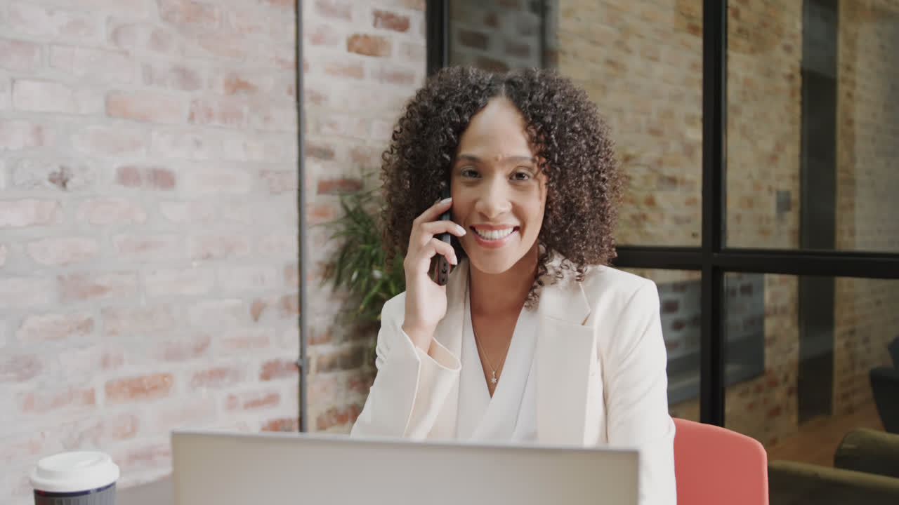 Retrato de empresária birracial feliz usando notebook e falando ao smartphone, em câmera lenta