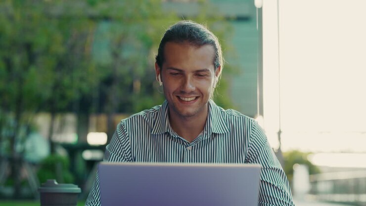 Young Man Working on Laptop Outdoors with Earbuds, Smiling