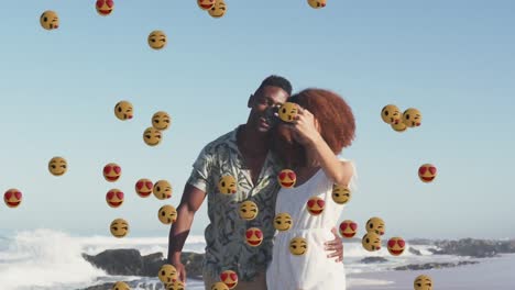 animation of smiling and love emojis over african american couple taking selfies on sunny beach