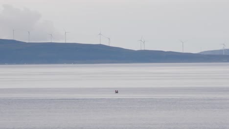 Plano-General-Estático-De-Un-Pescador-En-Un-Barco-Pesquero-En-Un-Océano-Tranquilo-Y-Un-Paisaje-De-Colinas-Con-Molinos-De-Viento-Giratorios-En-El-Fondo