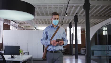 Caucasian-businessman-walking-through-corridor-wearing-facemask-holding-tablet