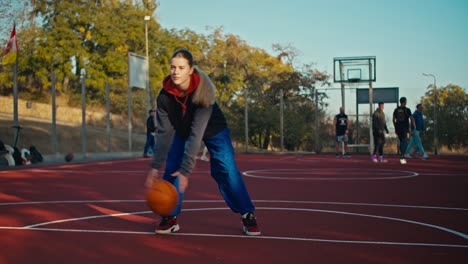 A-blonde-girl-in-a-sports-uniform-hits-an-orange-ball-from-the-floor-during-her-training-on-a-red-street-court-in-the-summer-morning.-Blonde-girl-in-sportswear-in-the-morning-for-basketball-training-on-the-street-court