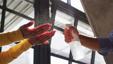 Mixed-race-male-cafe-worker-wearing-face-mask-disinfecting-hands-of-female-coworker