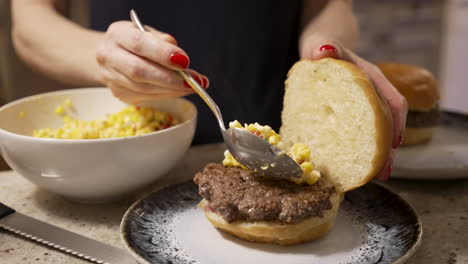 A-woman-spoons-corn-kernel-slaw-onto-freshly-grilled-burgers-on-a-counter-top