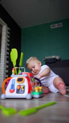 focused caucasian infant baby in white bodysuit sits on the floor in the room. chubby kid plays with a toy in front of him. low angle view. vertical video.