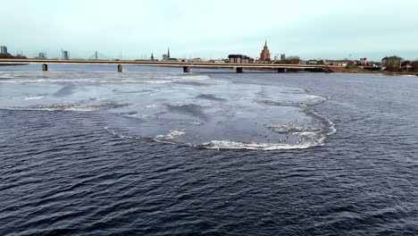 aerial, river daugava divides the european city of riga in half, in front you can see a piece of ice with many seagulls drifting on the piece of ice, traffic on the bridge