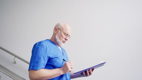 Un-Médico-Jefe-Seguro-De-Sí-Mismo,-Con-Gafas,-Barba-Gris-Y-Uniforme-Azul,-Baja-Las-Escaleras-Y-Anota-Cosas-Importantes-En-Su-Tableta-En-Una-Clínica-Moderna.-Un-Médico-Experimentado-Y-Seguro-De-Sí-Mismo-Anota-Los-Resultados-De-Sus-Exámenes-En-Una-Tableta.