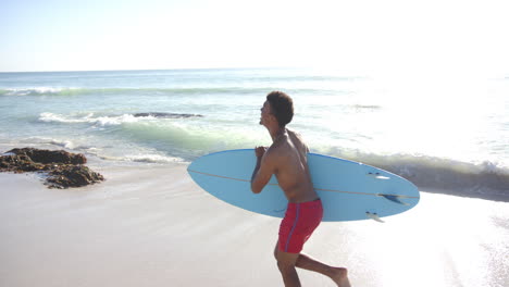 joven biracial lleva una tabla de surf azul claro en una playa soleada