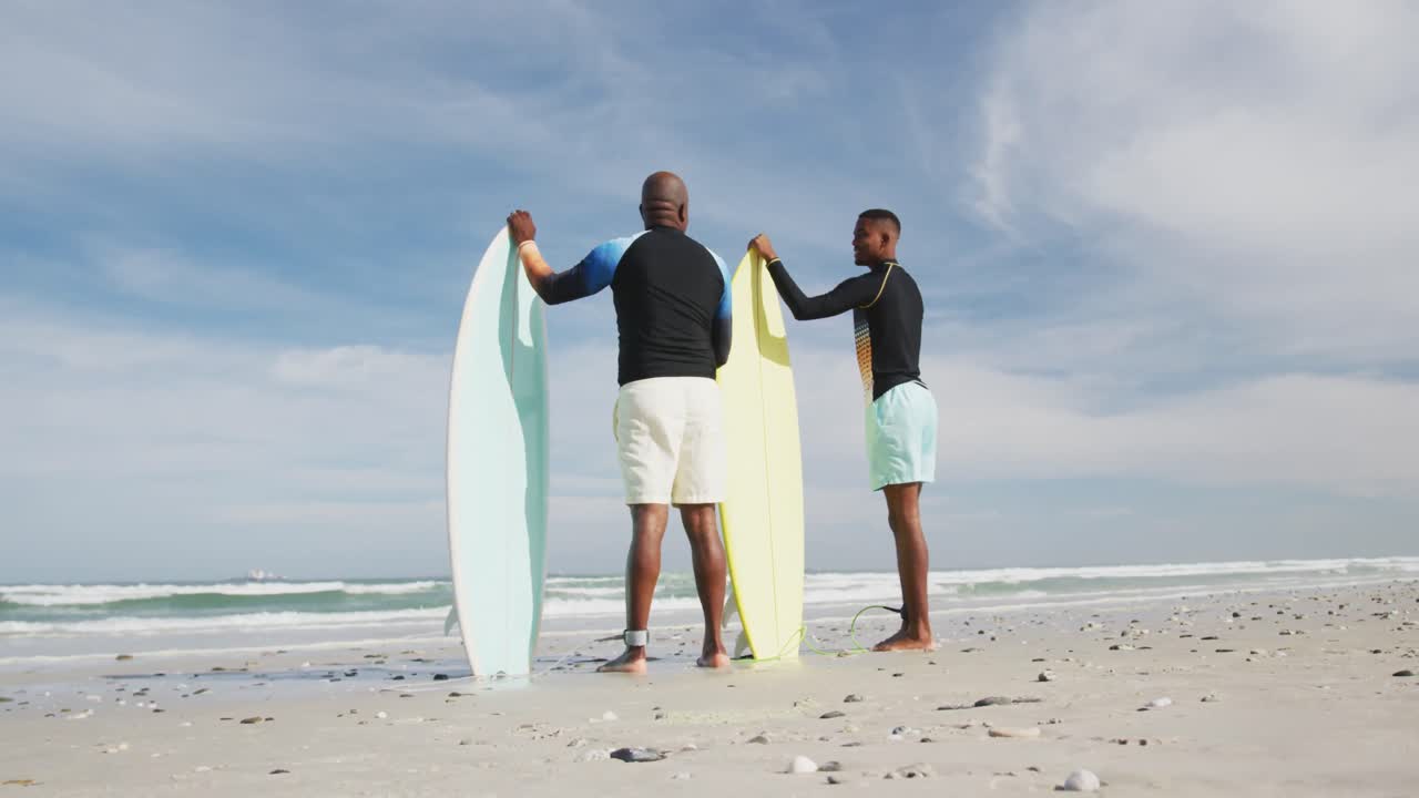 African american father and teenage son standing on beach holding surfboards and talking