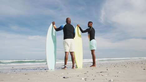 padre afroamericano y hijo adolescente de pie en la playa sosteniendo tablas de surf y hablando