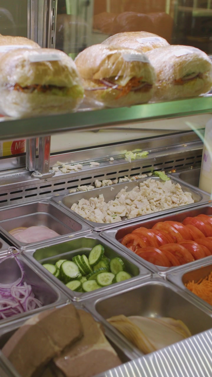 La telecamera scorre su panini da panetteria e ingredienti freschi assortiti per insalata, sotto una luce brillante.