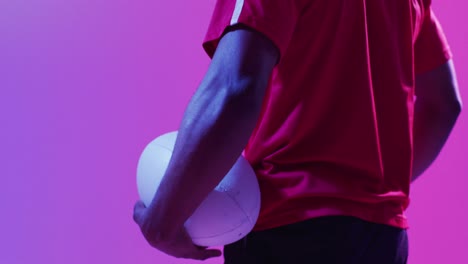 african american male rugby player with rugby ball over pink lighting