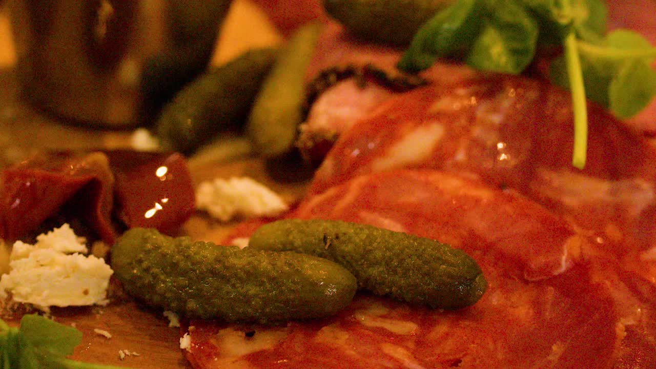 Close-up of fork picking up gherkin from assorted cold cuts, cheese, and pickles, warm lighting