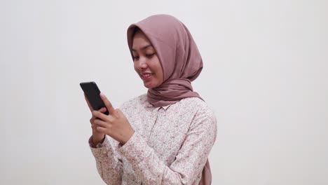 happy young asian muslim woman standing while using her cell phone and looking smiling at the camera