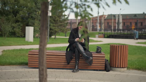 woman sitting on park bench using smartphone, dressed in black coat and boots, surrounded by lush green trees and a relaxed outdoor atmosphere in urban park setting