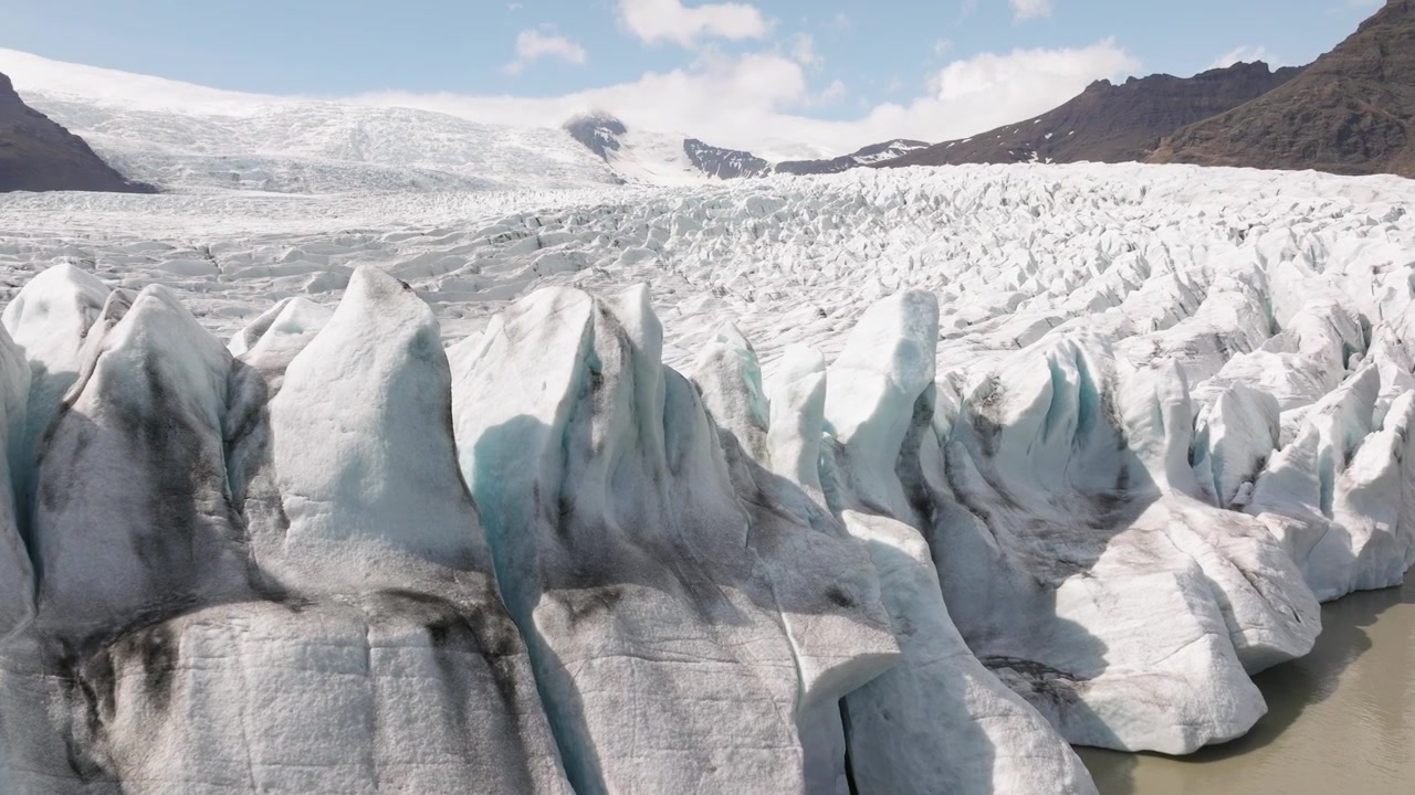Detaljert utsikt over Fjallsjökull-breens skarpe isformasjoner som fosser ned mot Fjallsárlón-lagunen på Island, innrammet av robuste fjell og en klar himmel