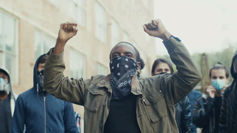 hombre afroamericano con bufanda en la cara y gritando con los brazos arriba en una protesta con un grupo multiétnico de personas en la calle