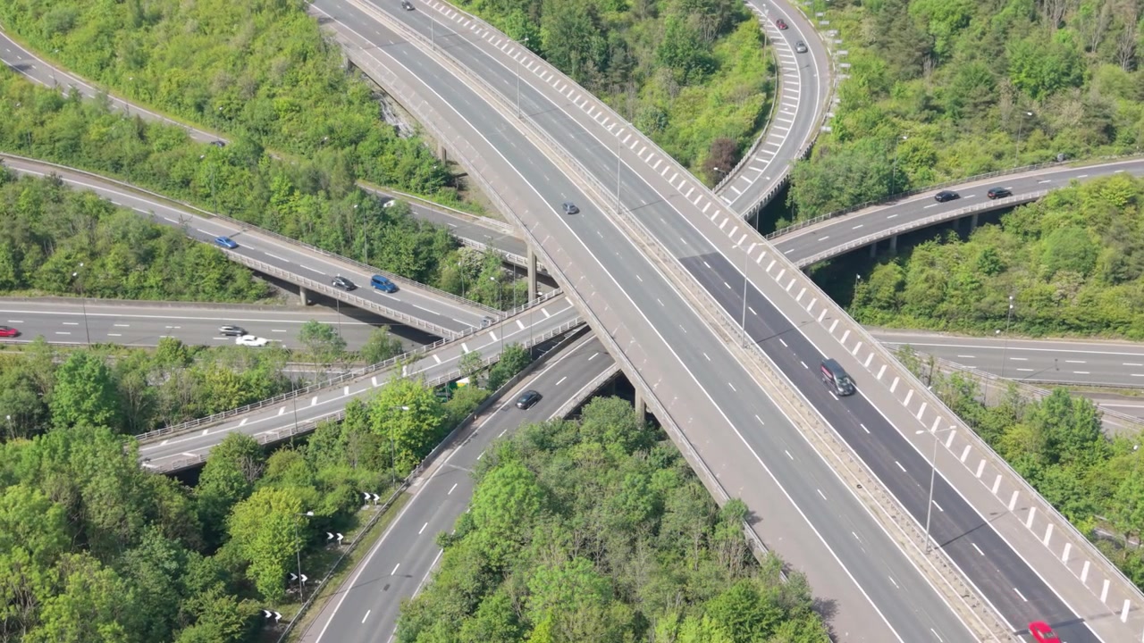 Aerial perspective of a multi-level highway interchange with many cars moving on roads and bridges. Dense green trees surround the busy junction. Traffic flows smoothly under a bright daytime sky.
