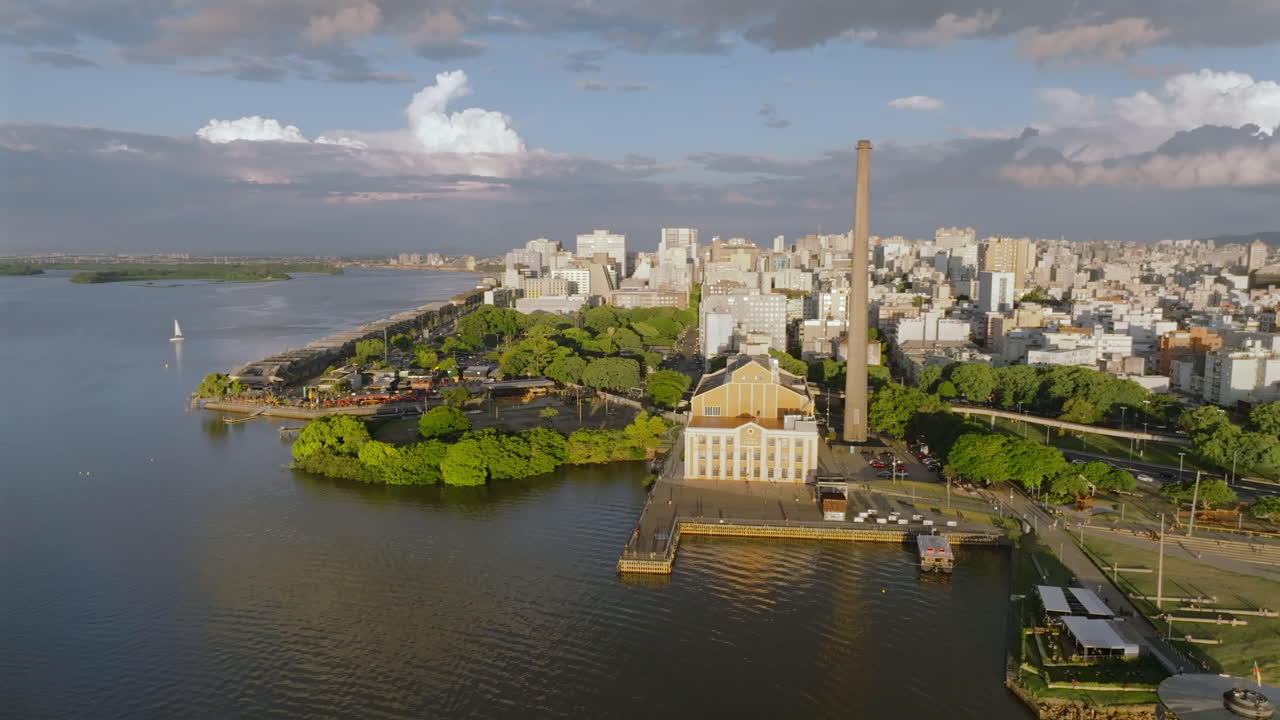 Drone aereo in volo laterale sopra il lago Guaiba che rivela lo skyline della città di Porto Alegre, il Gasometro e una barca a vela durante il tramonto. L'alta risoluzione cattura uno stormo di uccelli migratori che volano in formazione a V.