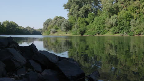 Trees-are-reflected-in-the-river's-rippling-water-with-foreground-rocks