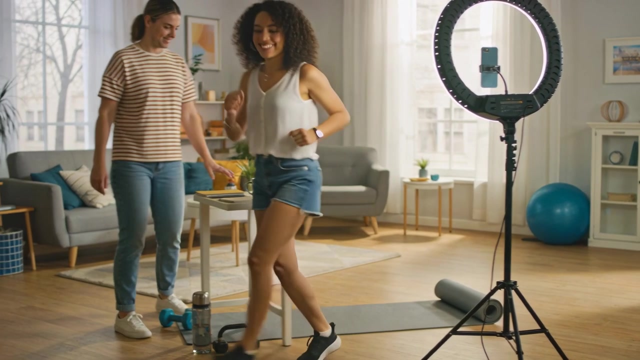 Starting curly woman in shorts leading dance steps on living room mat, partner stepping, ring light