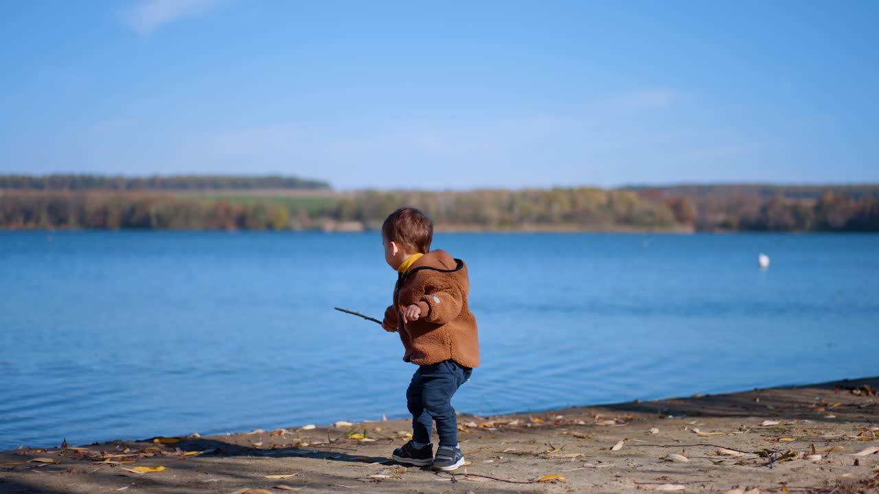 Little Caucasian kid is on the river bank. Baby boy puts the stick into water. Sunny autumn day in nature.