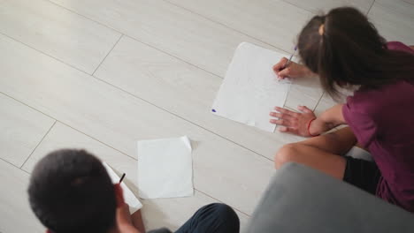 top down view of kids seated on floor drawing and learning on white papers with focus and creativity one girl with red rope around wrist sketching cat while boy on other side practices pencil drawing