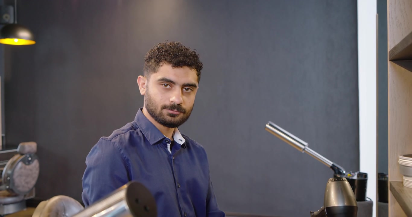 Estilista masculino de pie en el mostrador del salón, alcanzando y ajustando el secador y las máquinas de cortar pelo, preparándose para un cliente.