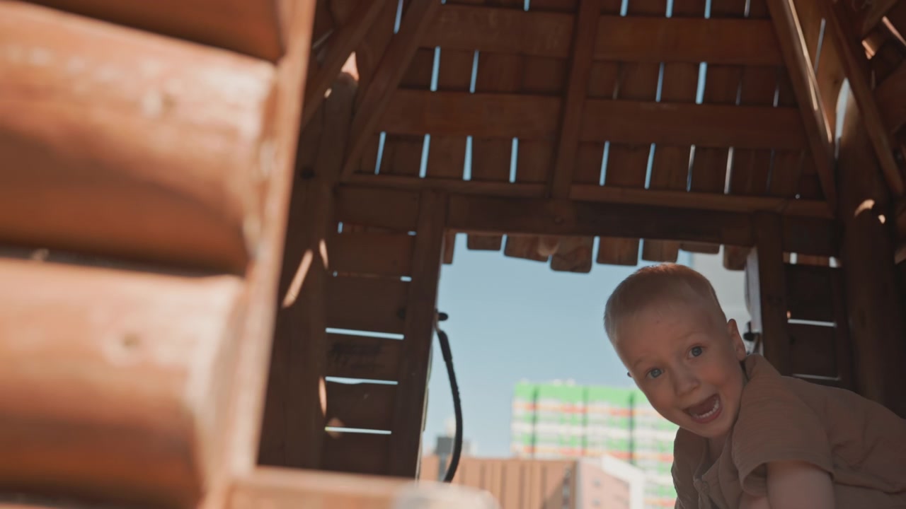 Young boy explores wooden playhouse moving through structure and looking out towards urban skyline. scene features warm sunlight streaming into rustic wood interior as boy interacts with his