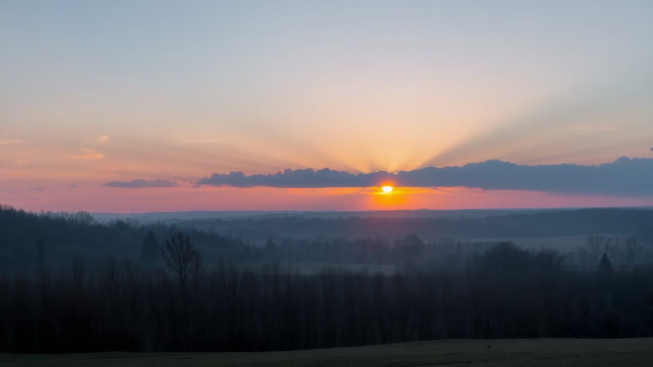 Auftauchende Sonne bricht durch eine niedrige Wolkenbank über einem grasbewachsenen Hang, beleuchtet ein nebliges Tal.