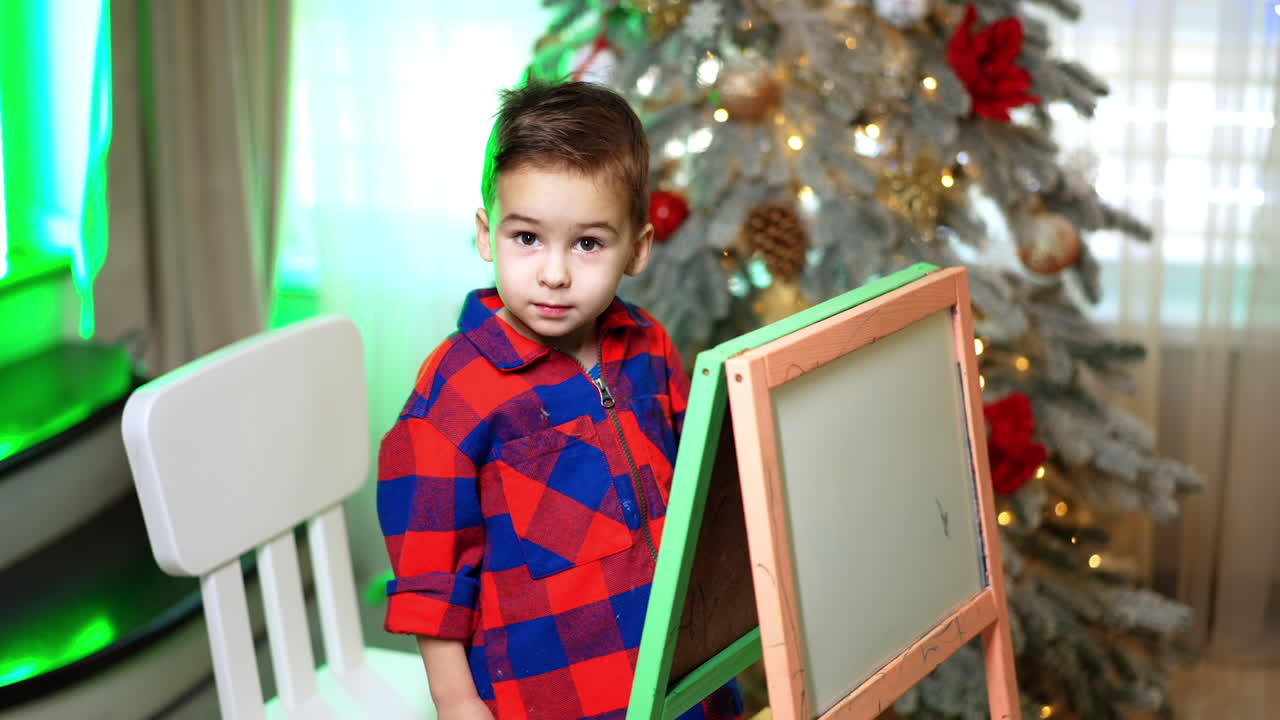 Kid in checkered shirt draws on the blackboard. Beautiful Christmas tree at backdrop