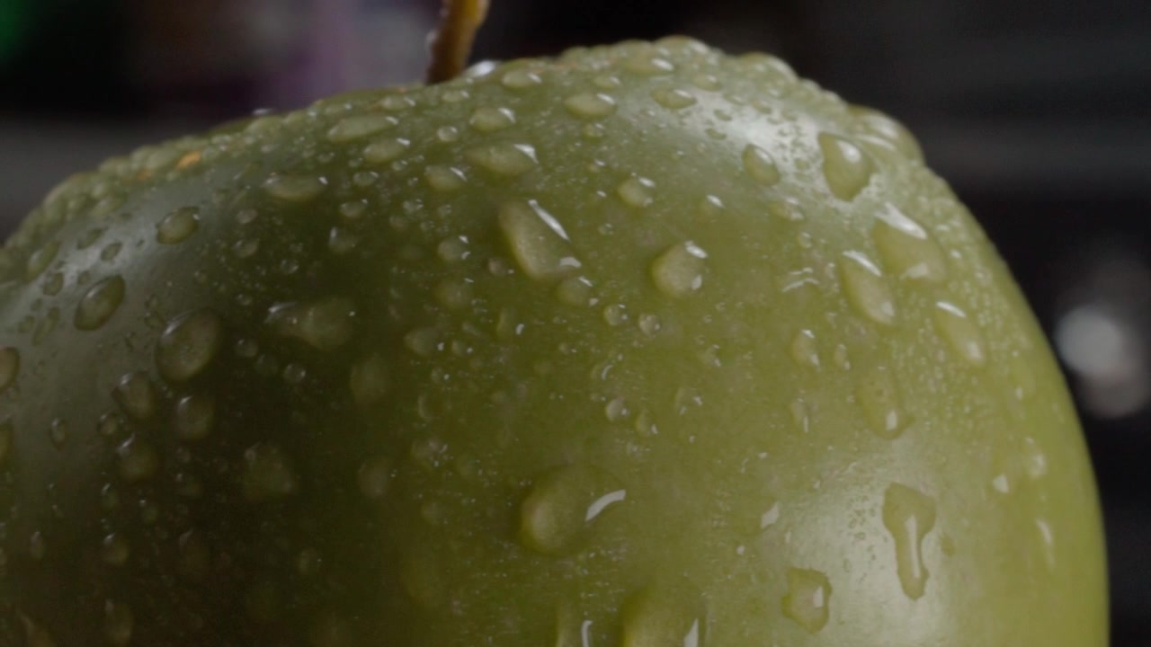 Primo piano macro di mela verde coperta da goccioline d'acqua fresca sulla sua buccia. Texture dettagliata e lucentezza naturale della superficie sono visibili su tutto il frutto.