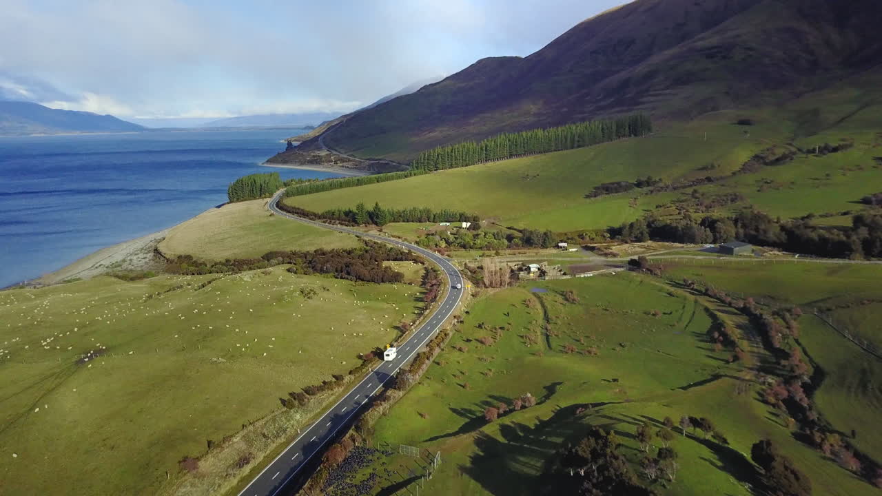 видеосъемка с воздуха: фургон едет по прибрежной дороге с зелеными лугами, затем панорамирование на голубой океан и снежные горы