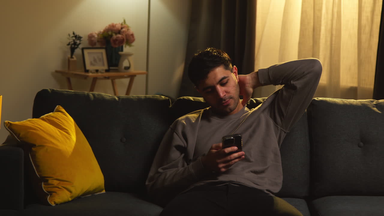 Young Man Spending Evening At Home Sitting On Sofa With Mobile Phone Scrolling Through Internet Or Social Media 6