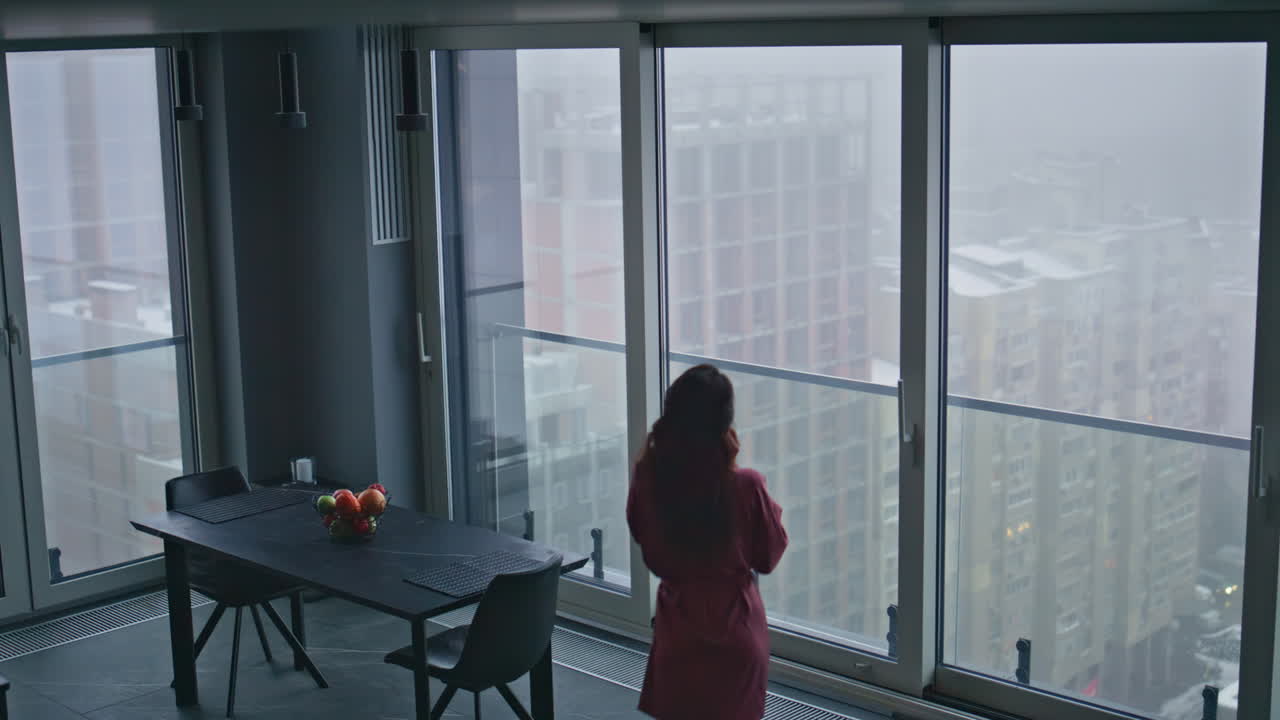 молодая домохозяйка звонит в смартфон, ходит босиком по панорамному окну в квартире.